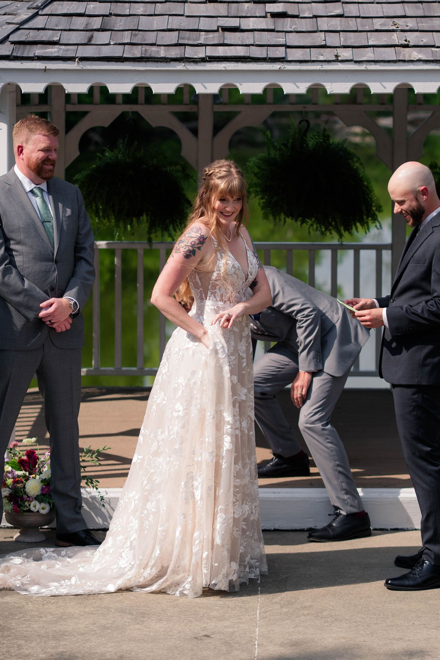 Bride in lace wedding gown laughing during outdoor ceremony near white gazebo with groom.