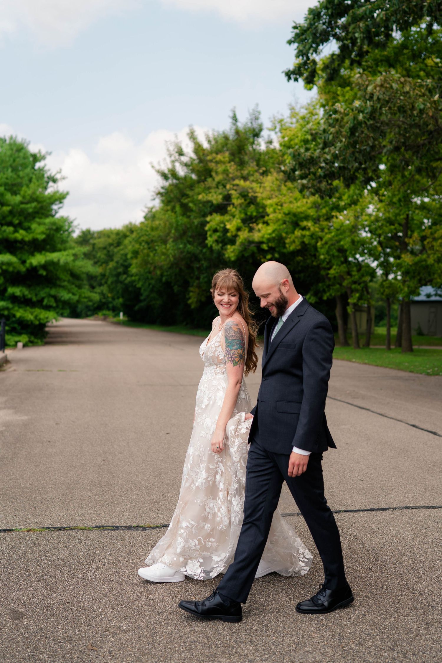 Bride and groom walking hand in hand down tree-lined path on wedding day.