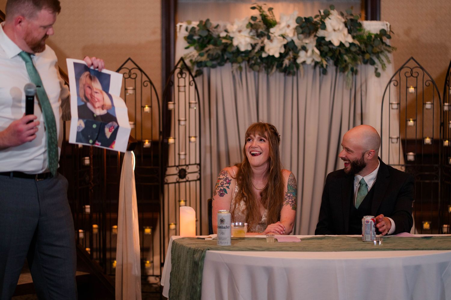 Best man holds up childhood photo during wedding reception speech as bride and groom laugh at table.