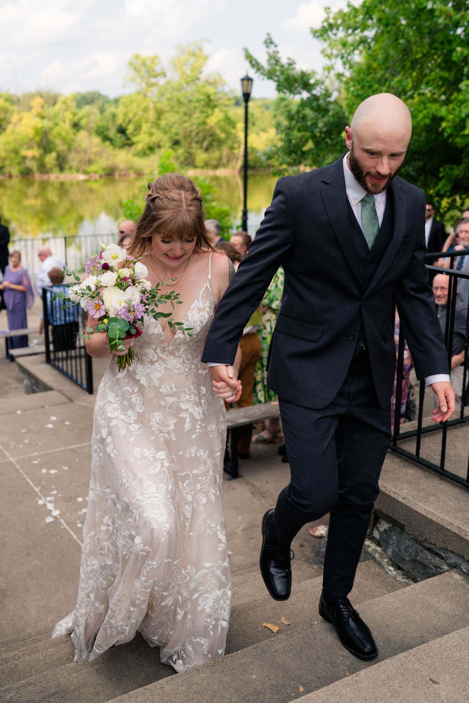 Newlywed couple walks hand in hand down stone steps after outdoor wedding ceremony near lake.