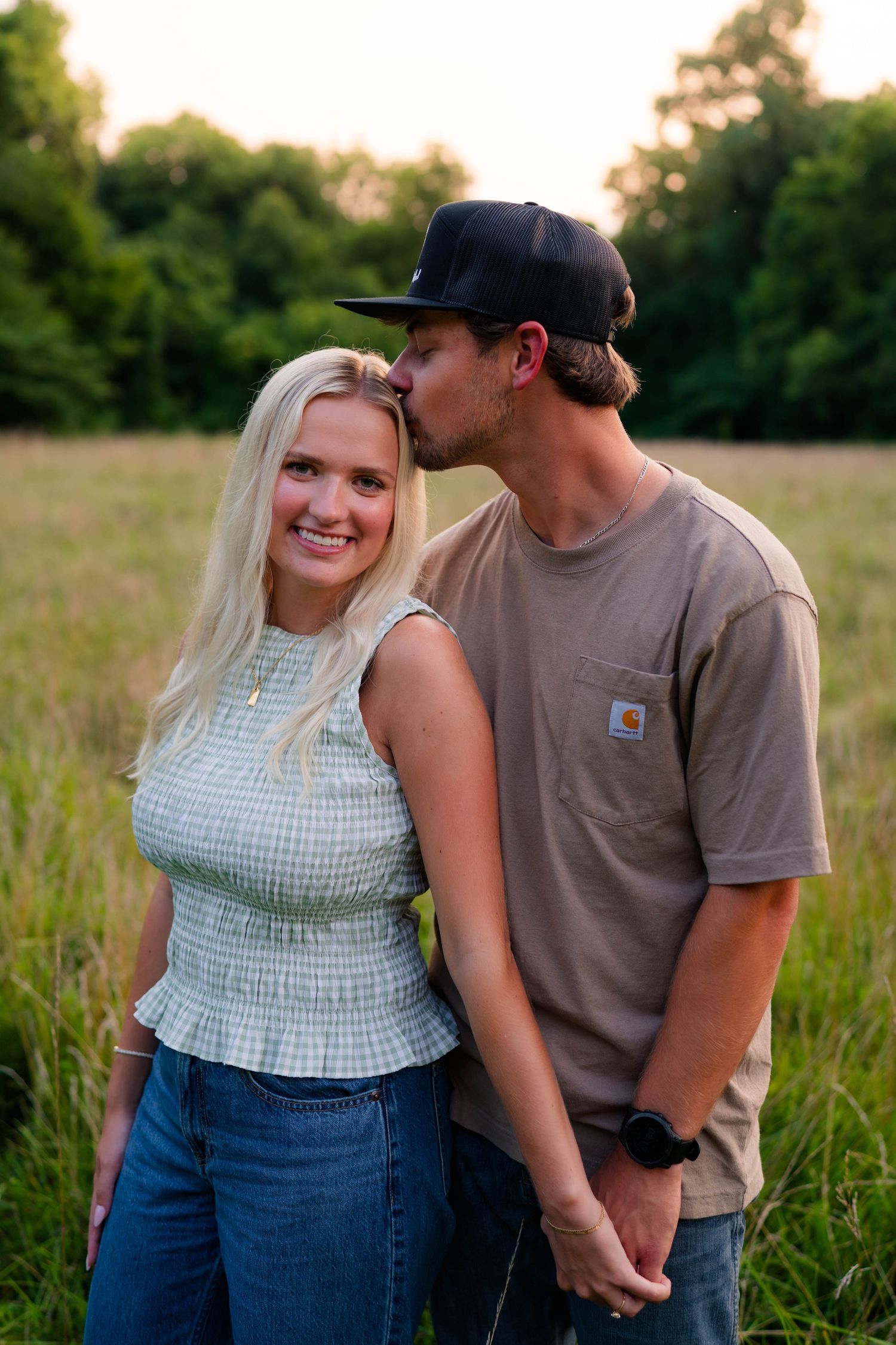 Couple posing in a green field at sunset, man kissing woman on the head while holding hands.