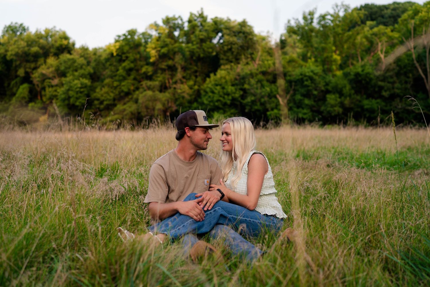 Couple sitting together in tall grass field, sharing a romantic moment at golden hour.