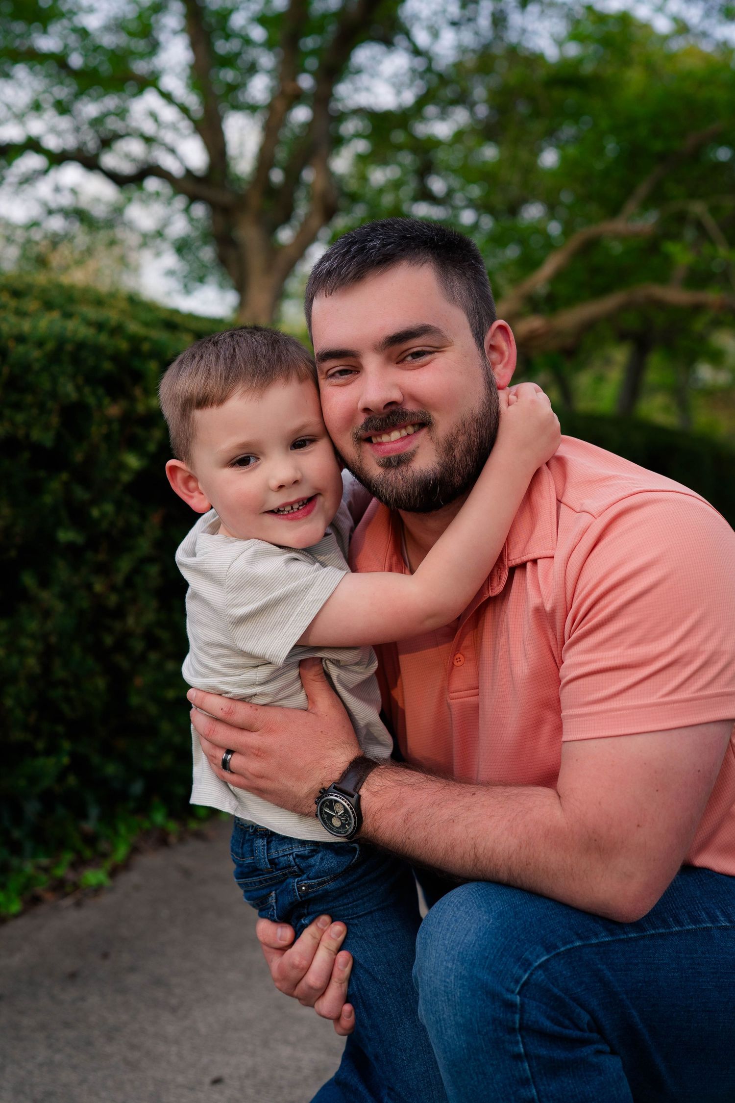 A smiling father crouches outdoors hugging his happy toddler son, both wearing casual clothes with green hedges behind them.