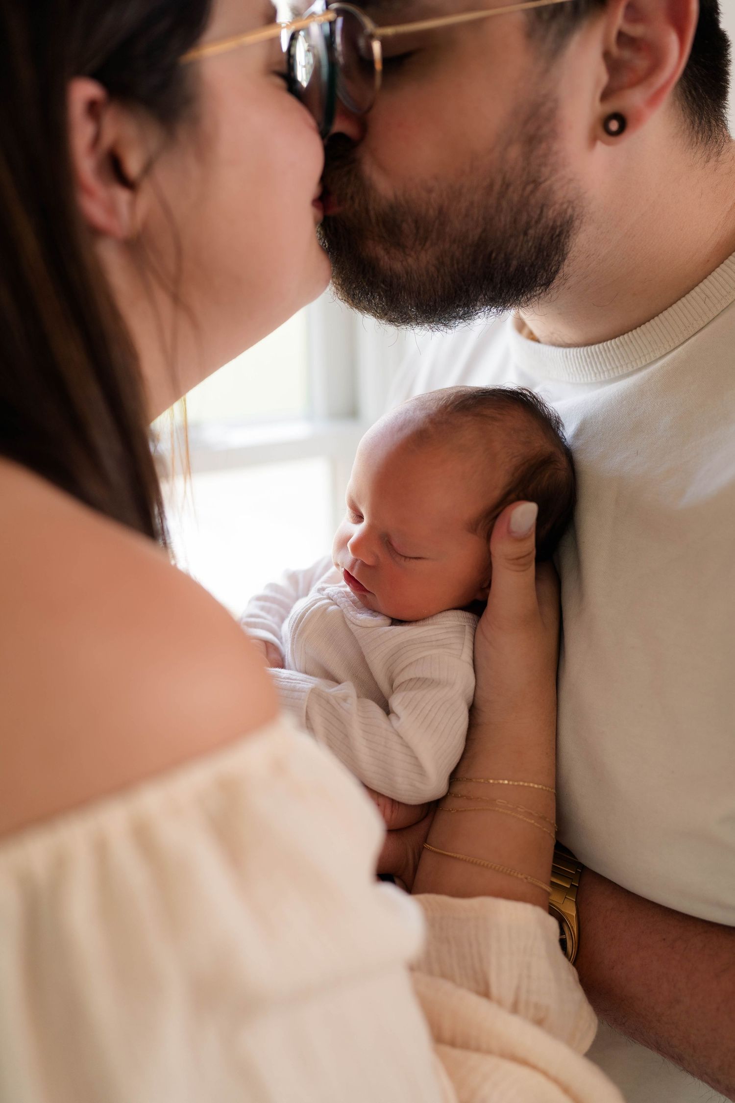 Parents share a kiss while holding their sleeping newborn baby together near a bright window.