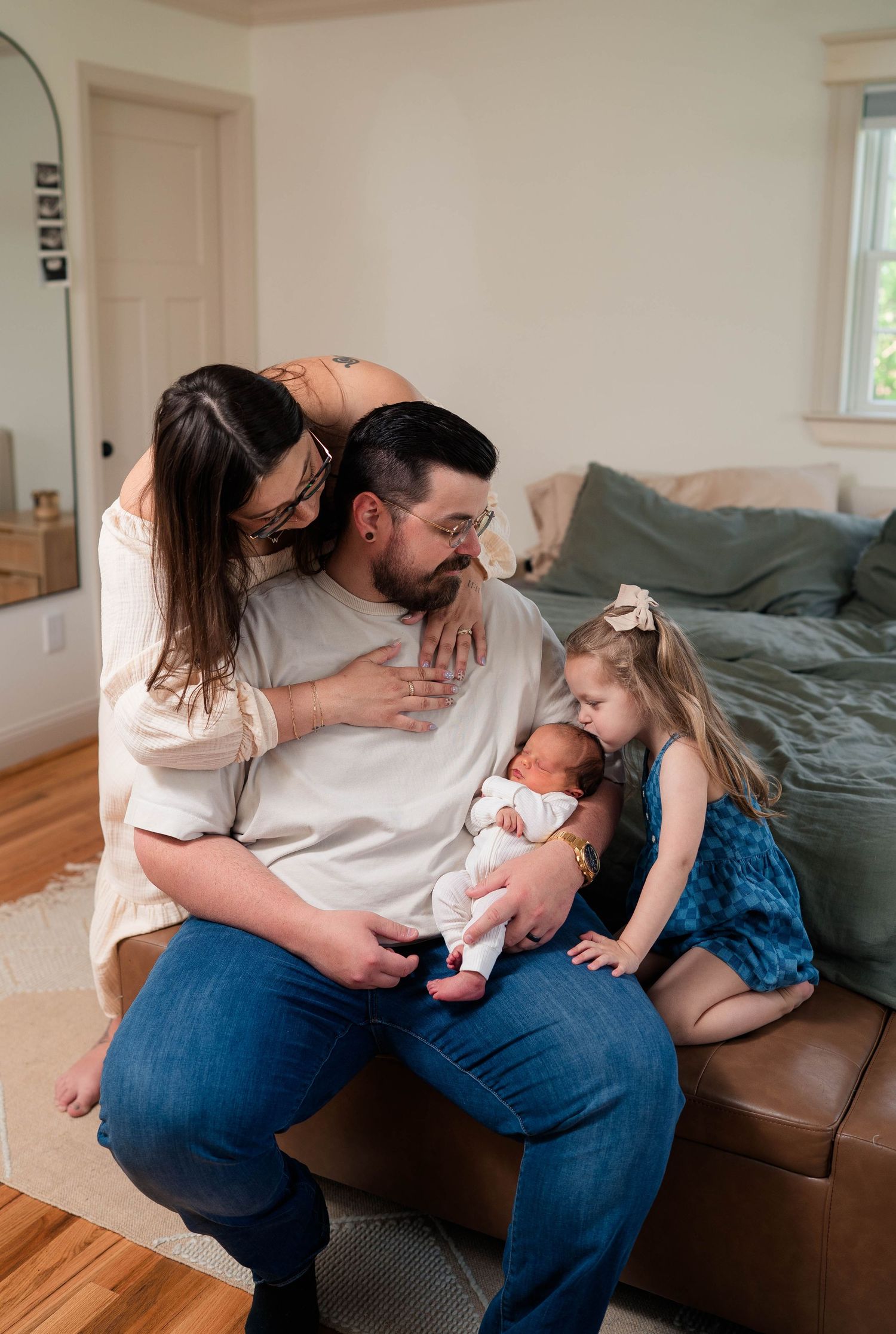Family of four shares a tender moment on a leather ottoman, dad holding newborn with toddler nearby.