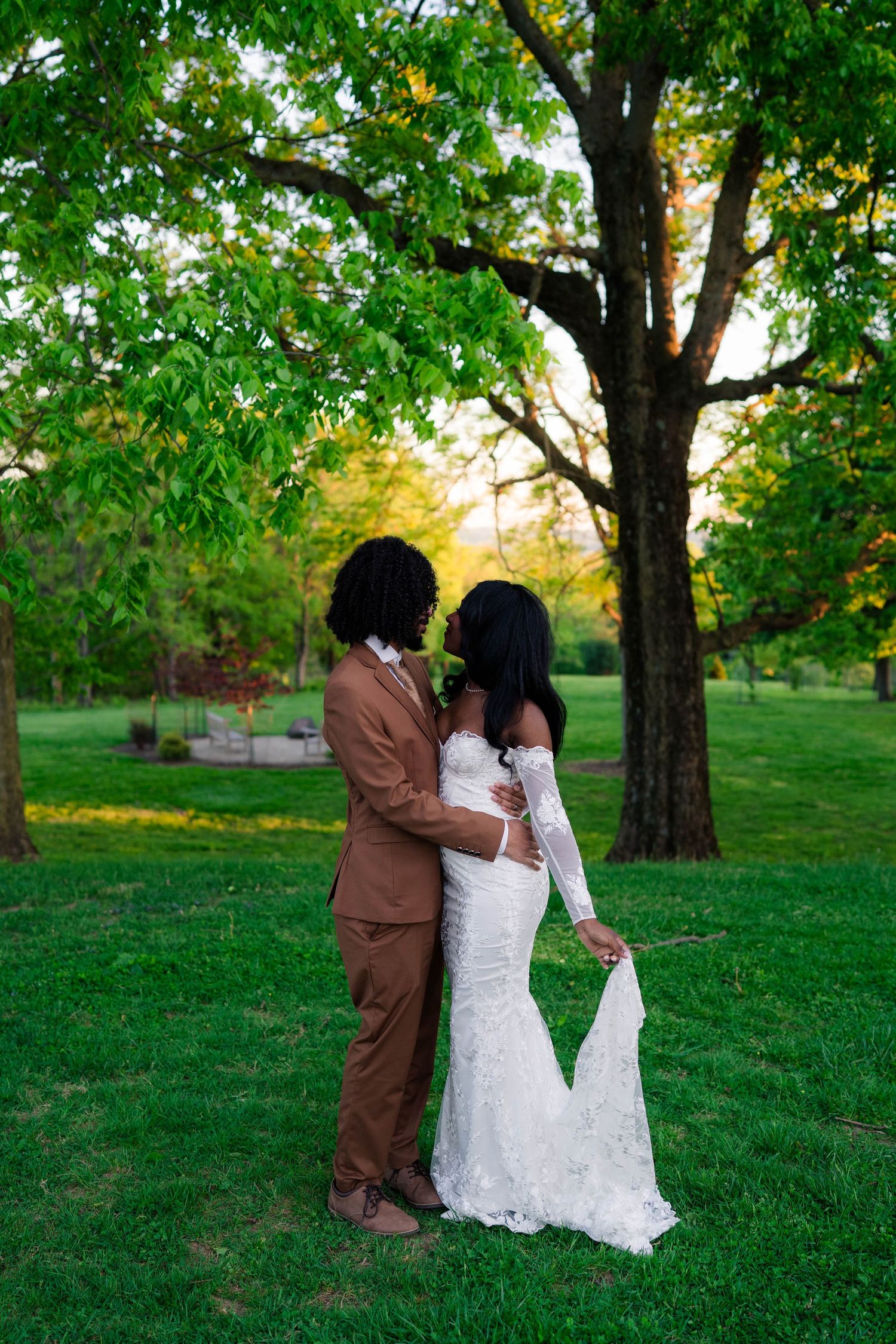 A Black couple in wedding attire embraces in a lush green park, bride in white lace gown, groom in tan suit, at golden hour.