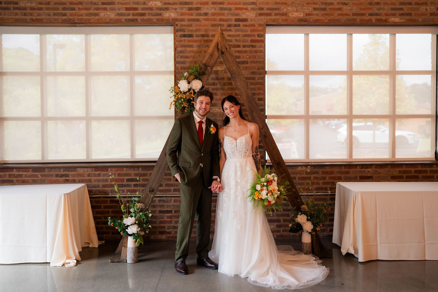 Bride and groom pose smiling before triangular wooden arch with floral arrangements at brick wall venue.