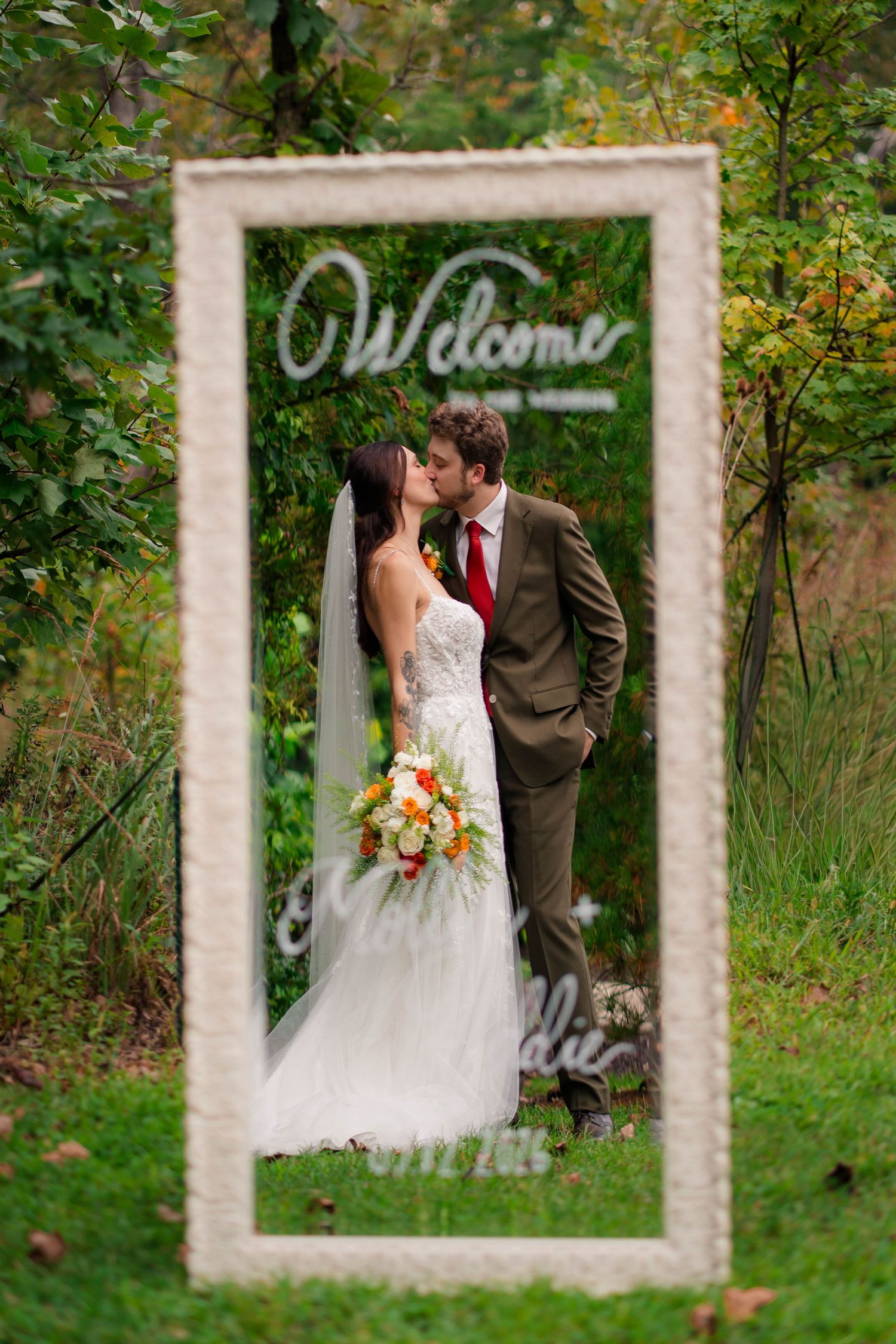 Bride and groom kissing framed by a decorative welcome mirror sign in an outdoor garden setting.