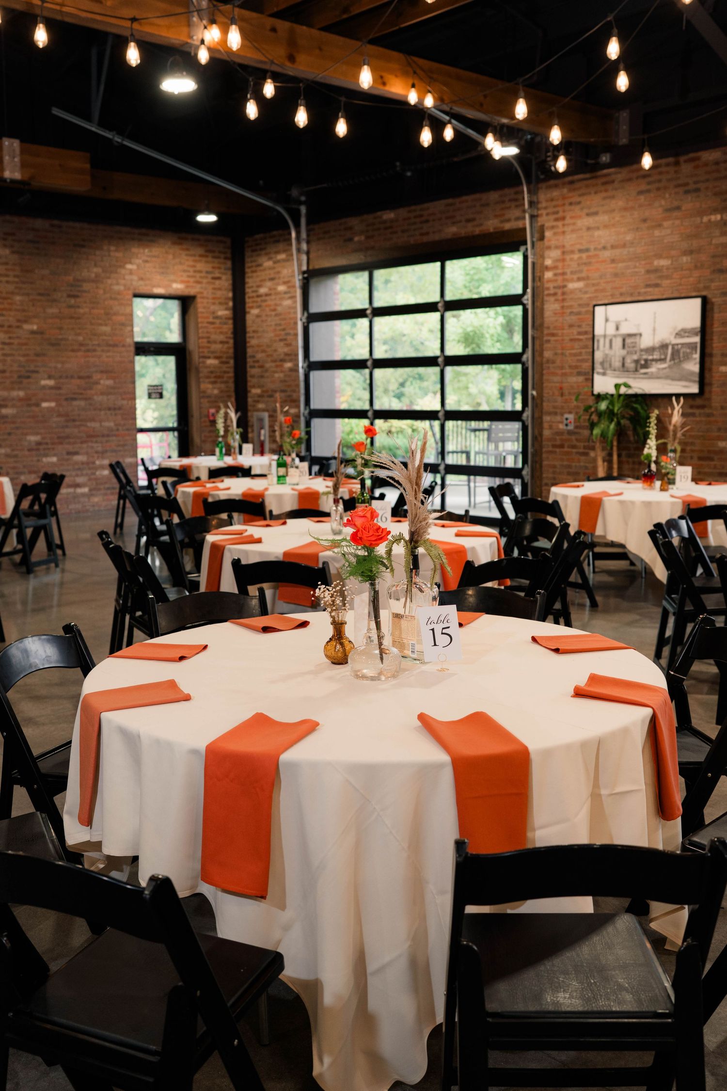 Wedding reception hall with round tables, orange napkins, black chairs, and Edison bulb string lights in brick venue.