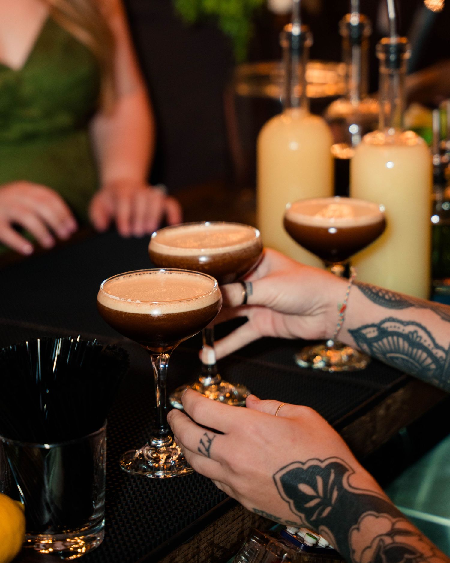 Tattooed hands holding espresso martinis at wedding reception bar with cream liqueur bottles.