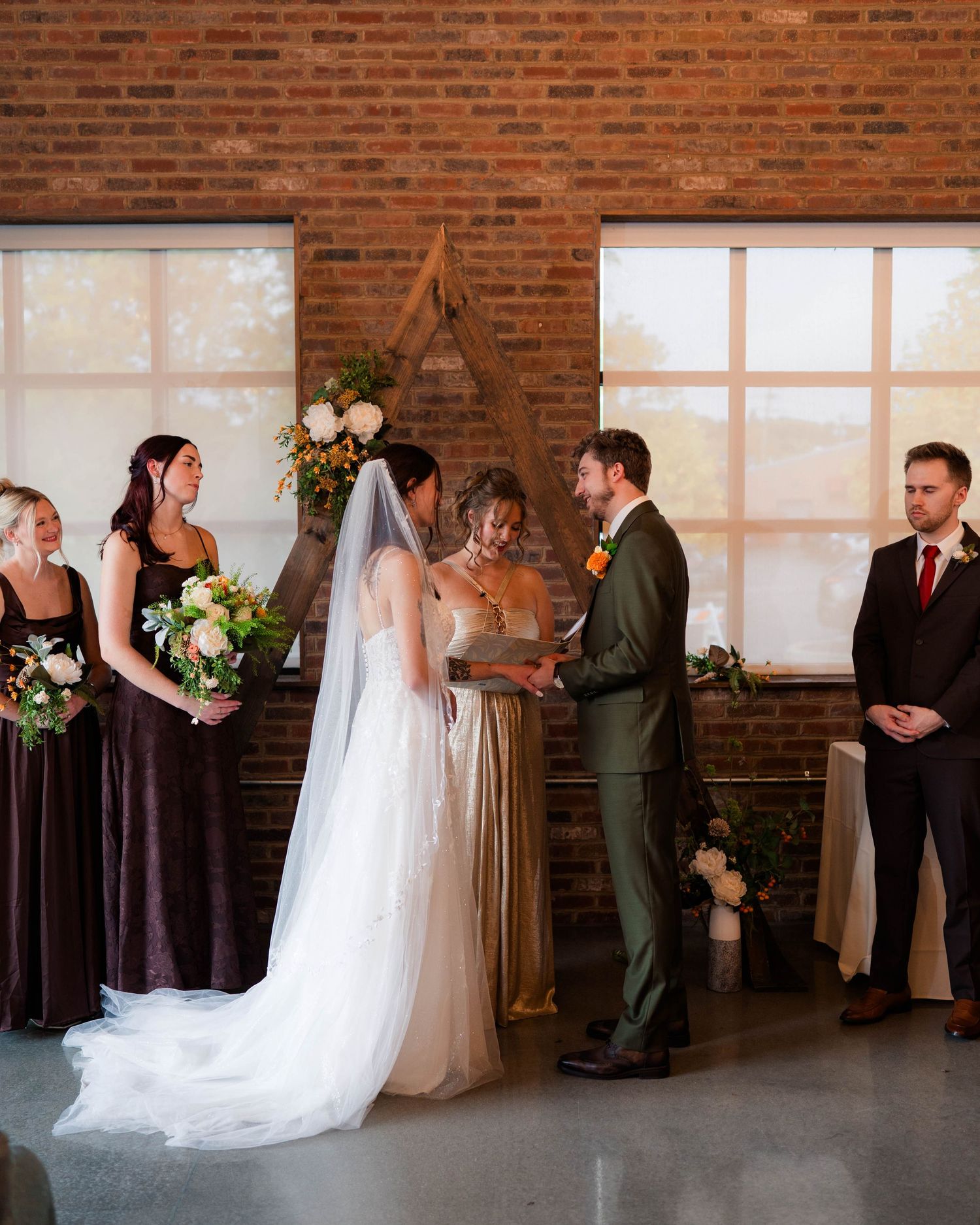 Bride and groom exchanging vows at altar with wedding party in rustic brick venue.