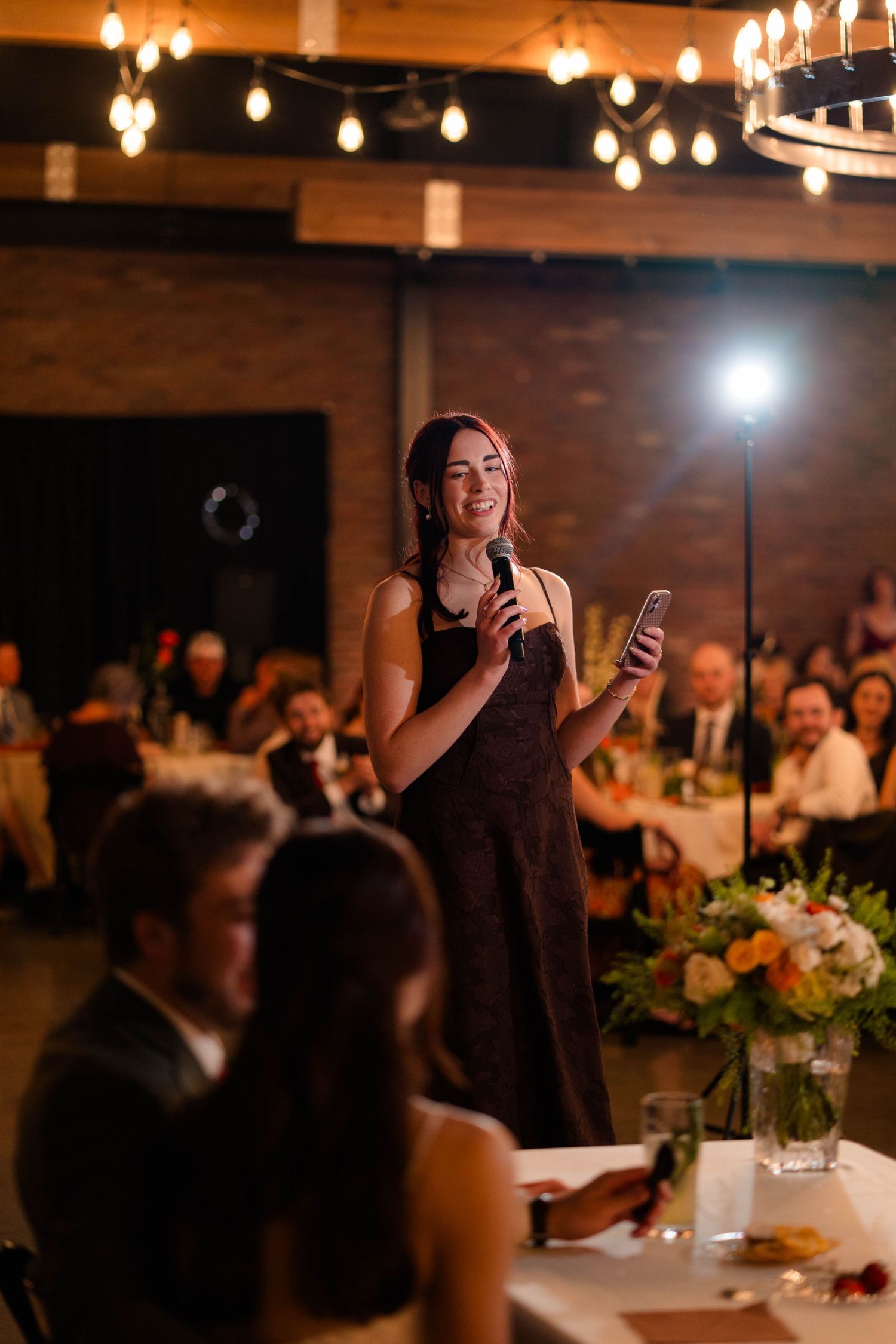 Woman in dark dress giving speech with microphone at warmly lit wedding reception venue.