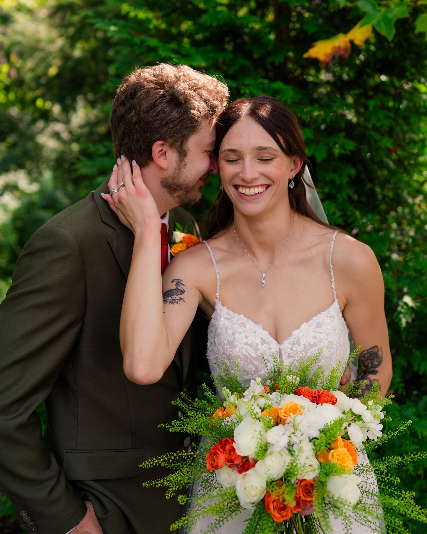 Groom in olive suit whispers to laughing bride holding white and orange bouquet outdoors amid greenery.