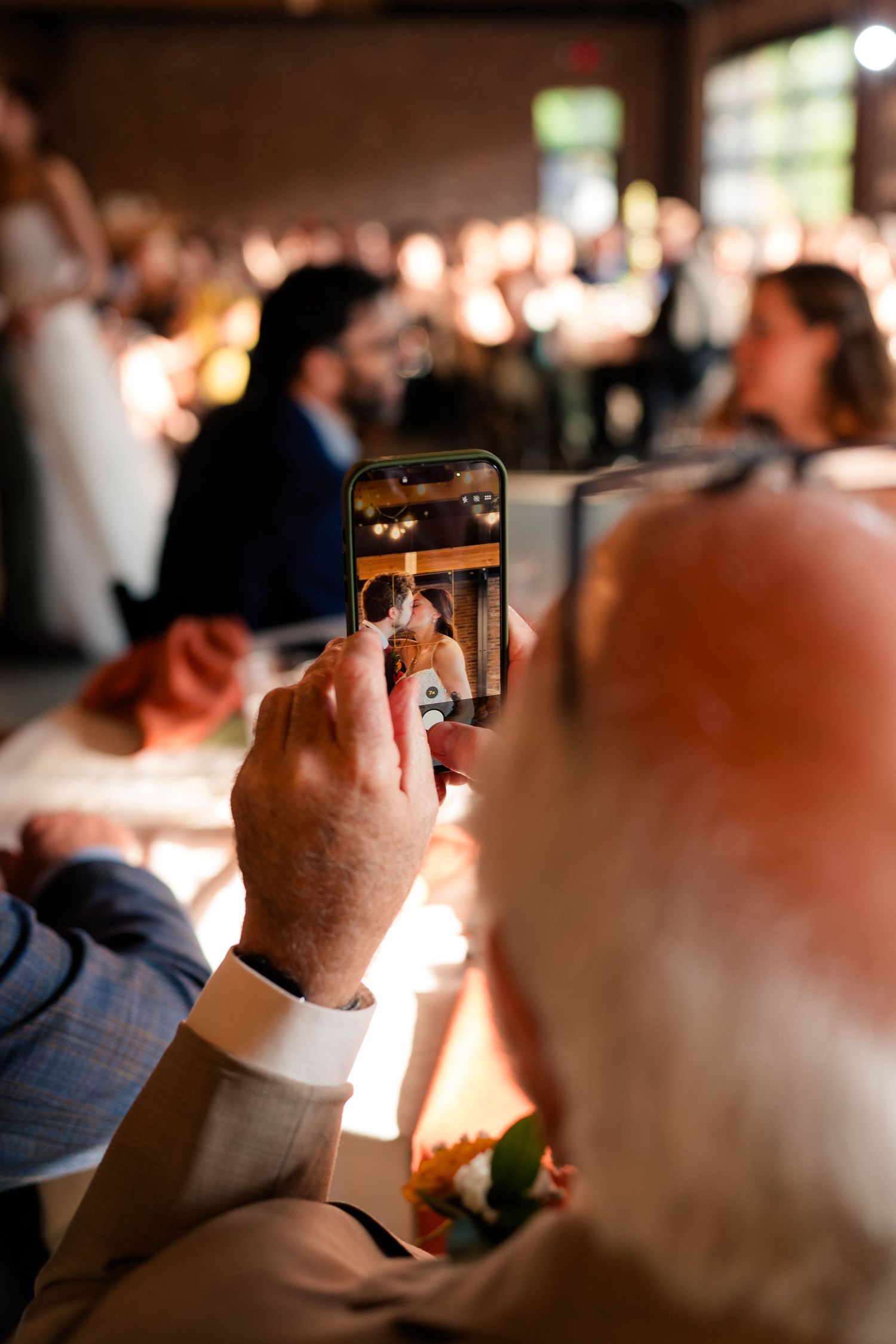 Wedding guest photographs the couple's first kiss on a smartphone during an indoor wedding reception.