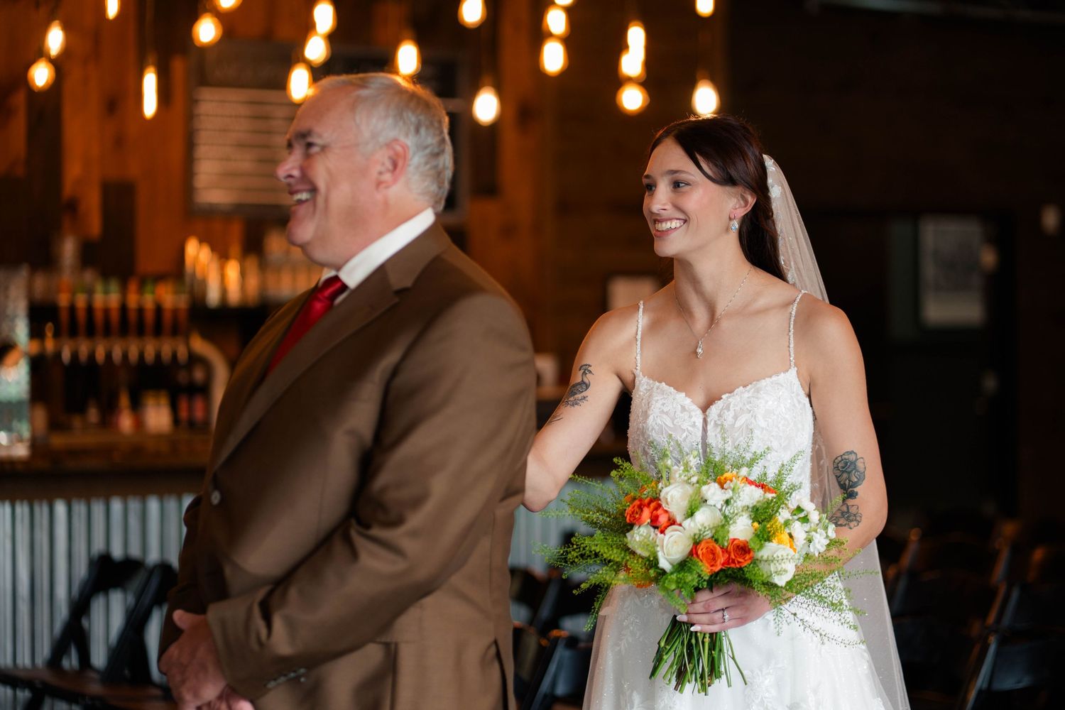 Bride walks down the aisle smiling, holding a colorful bouquet, accompanied by an older man in a brown suit.