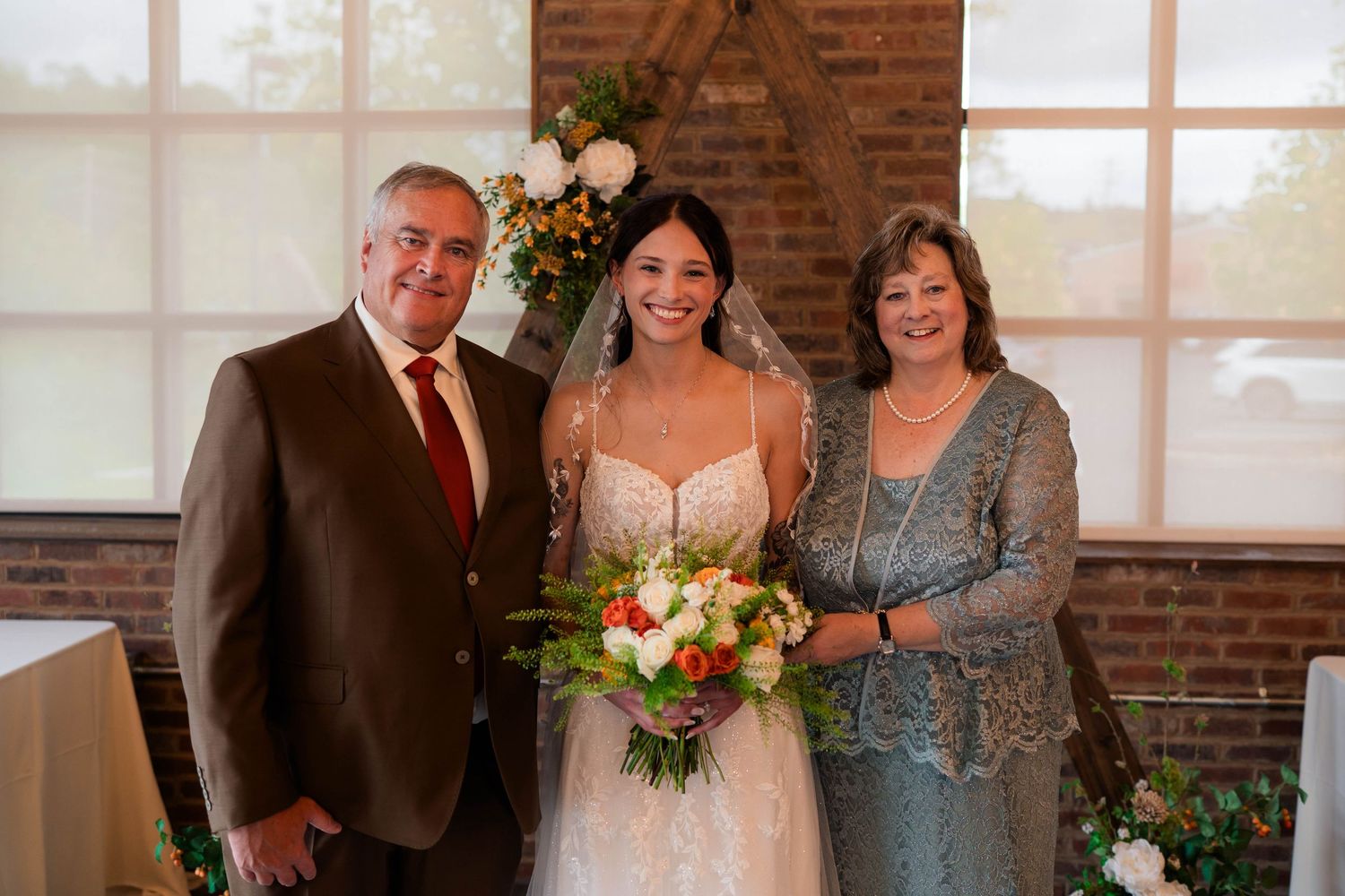 Bride with bouquet posing with parents in wedding venue decorated with floral arch.