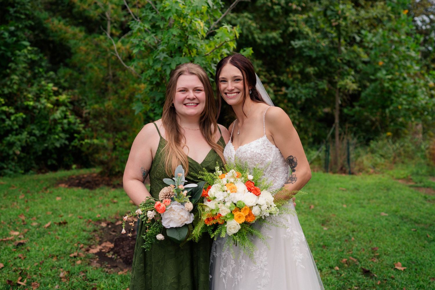 Bride in lace gown and bridesmaid in green dress smile together holding bouquets in garden setting.