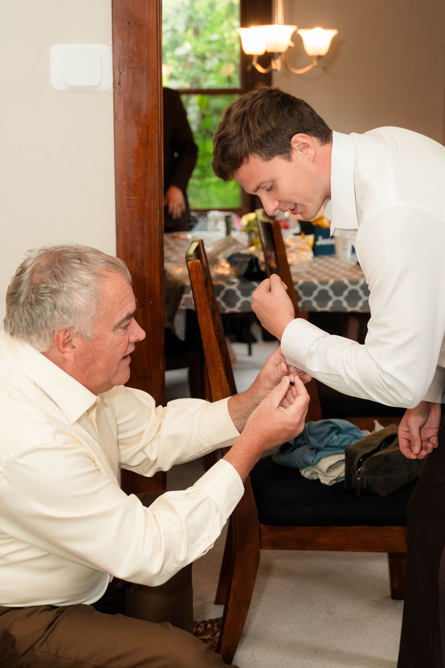 Older man helps young groom fasten cufflinks on his white dress shirt while smiling indoors.