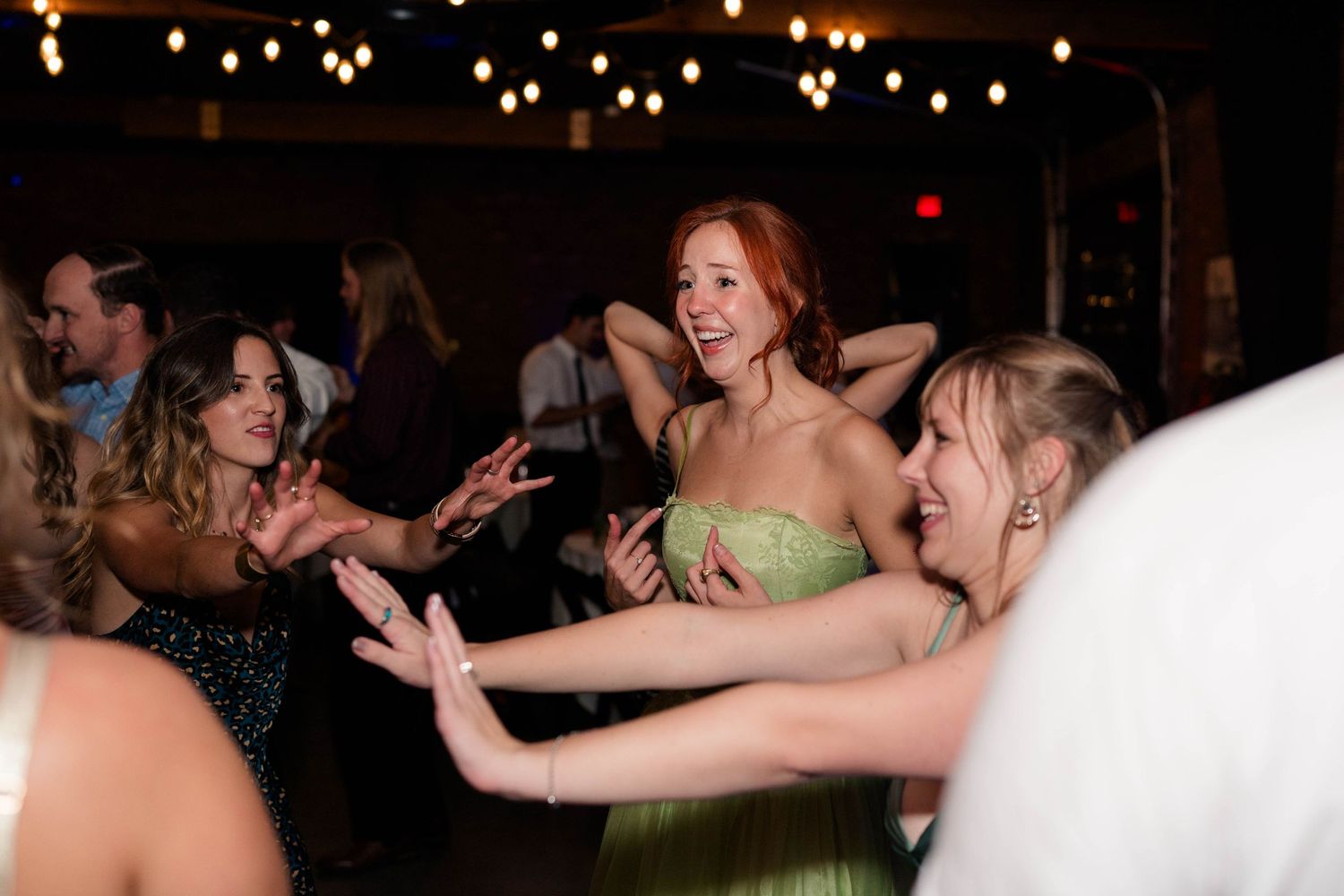 Bridesmaids in green dresses laugh and dance together joyfully on the wedding reception dance floor.