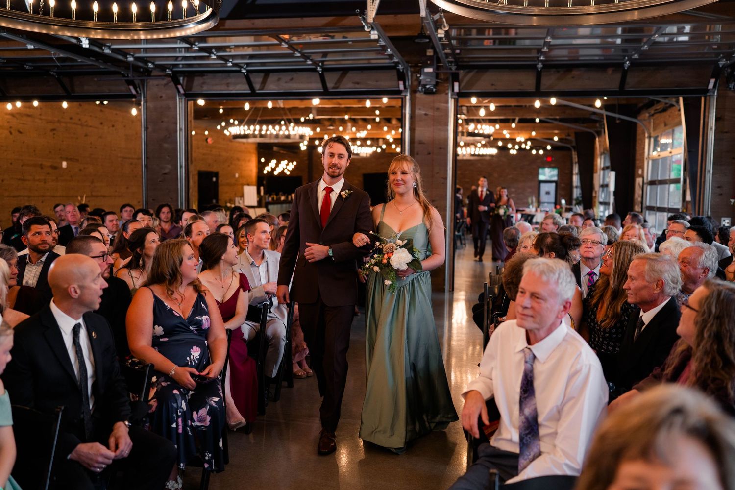 Bridesmaid in sage green gown walks down aisle with groomsman during packed indoor wedding ceremony.