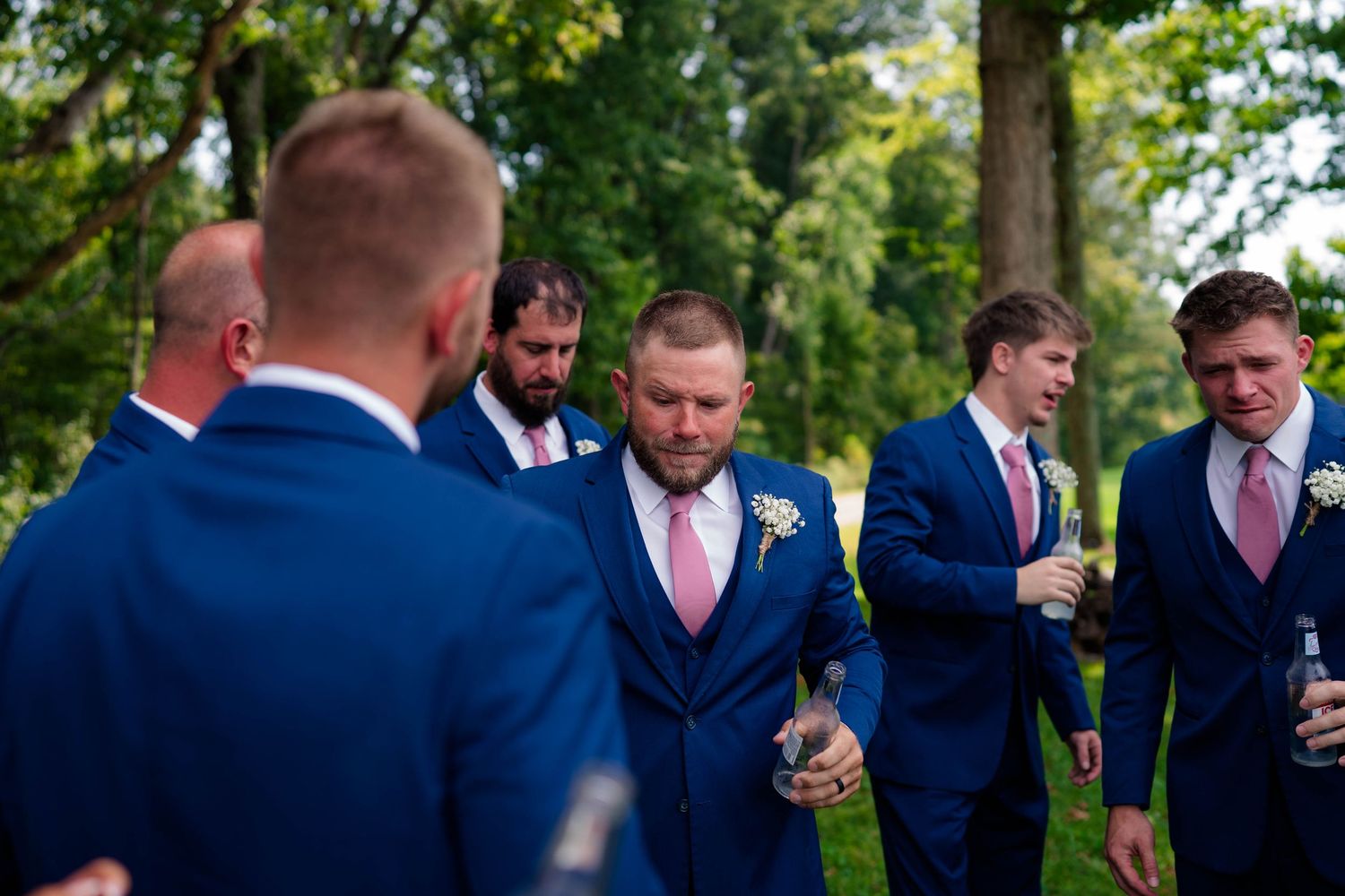 Groomsmen in navy blue suits with pink ties laughing and socializing outdoors before wedding ceremony.