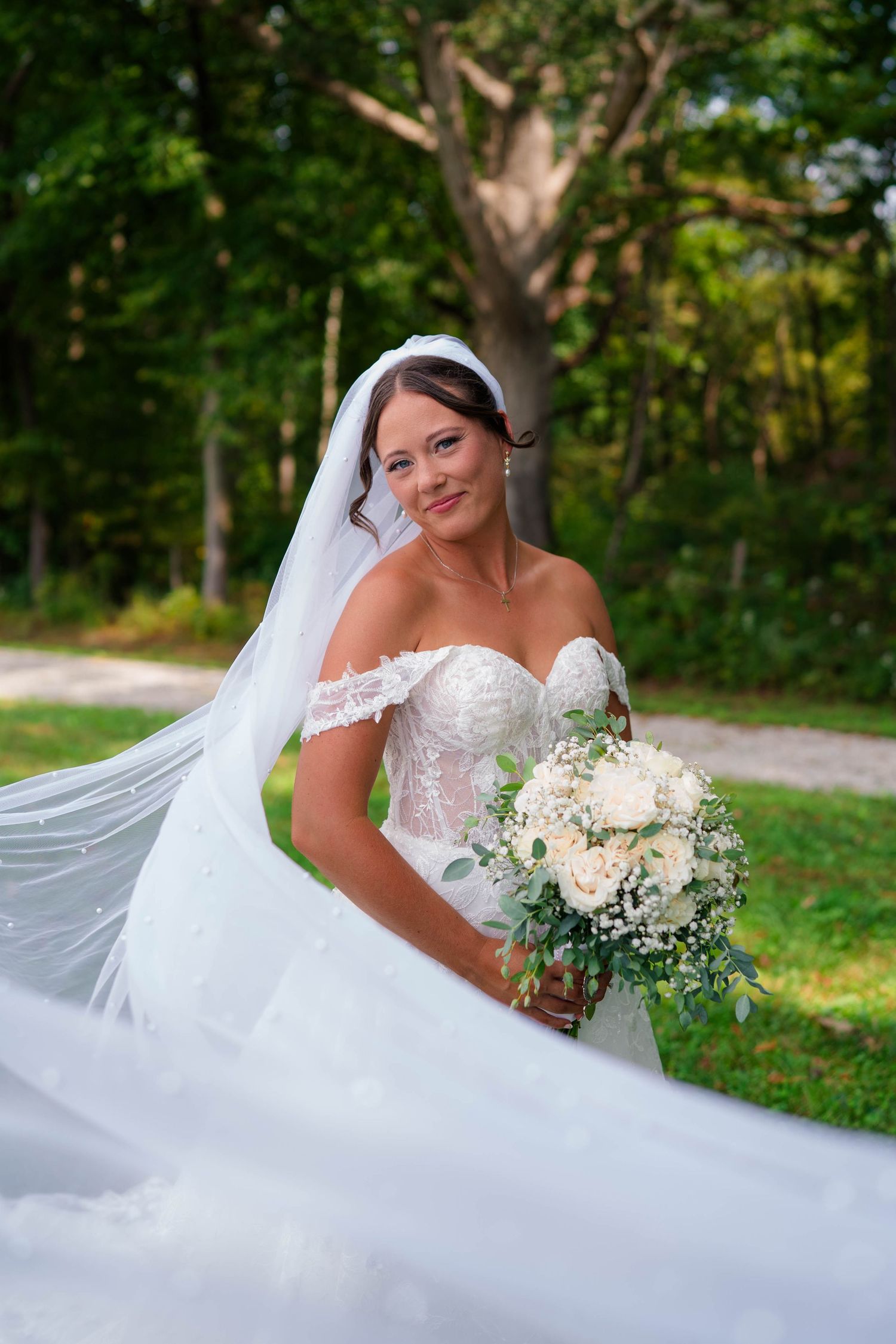 Bride in off-shoulder lace gown holds white bouquet with flowing veil in outdoor garden setting.