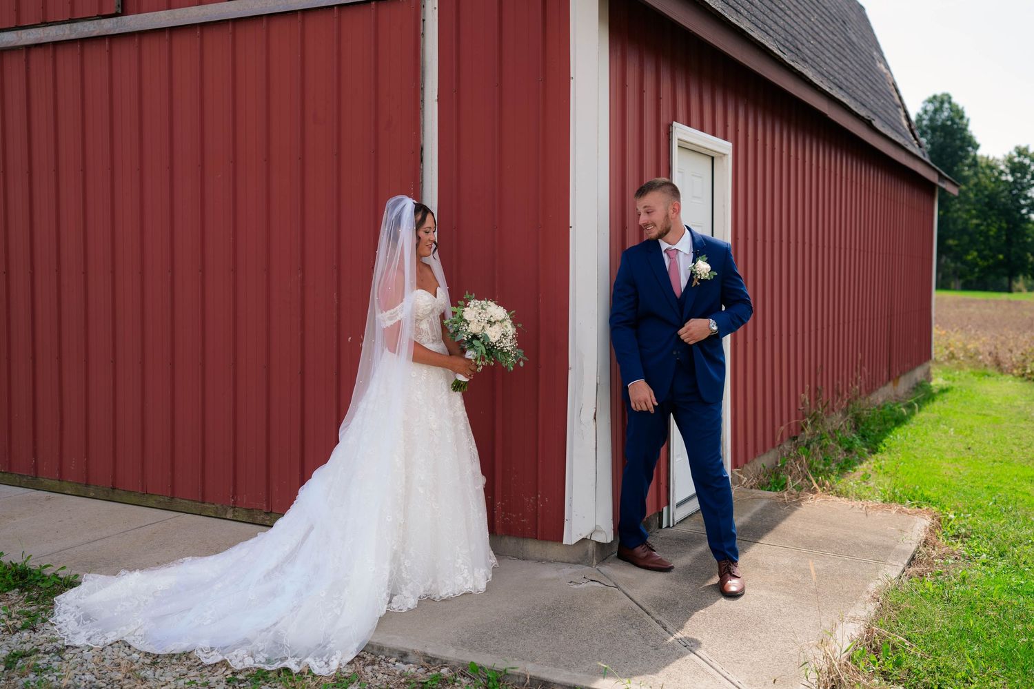 Groom turning to see bride in lace wedding dress during first look beside a red barn.
