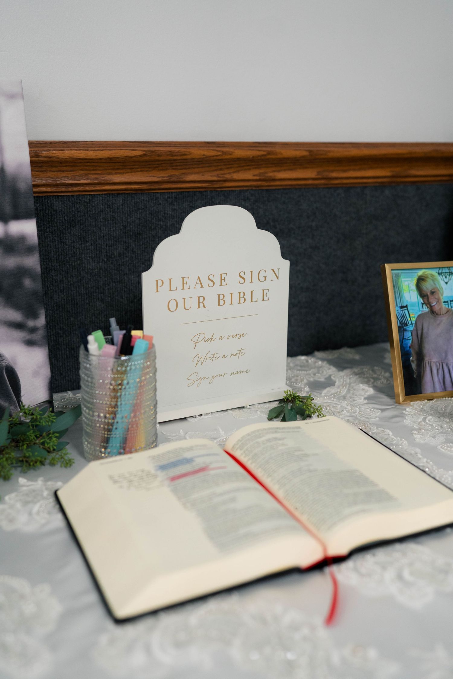 Open Bible guest book with colorful pens and a sign reading Please Sign Our Bible on a decorated table.