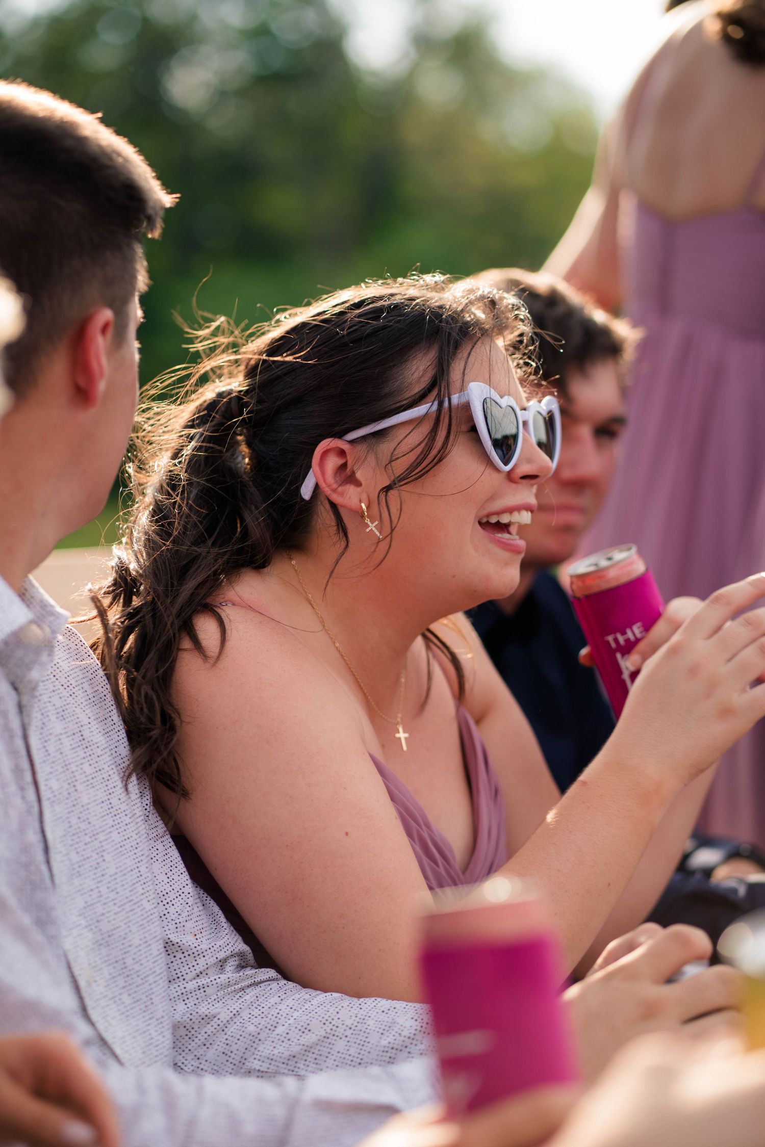 Smiling woman in heart sunglasses and lavender dress enjoys drinks at an outdoor wedding celebration.
