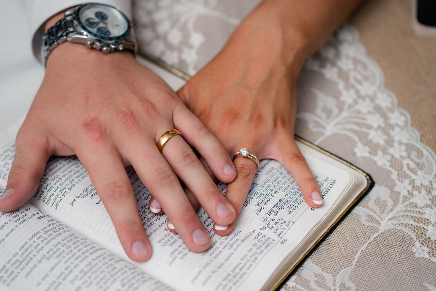 Bride and groom place hands on open Bible displaying their gold and diamond wedding rings.