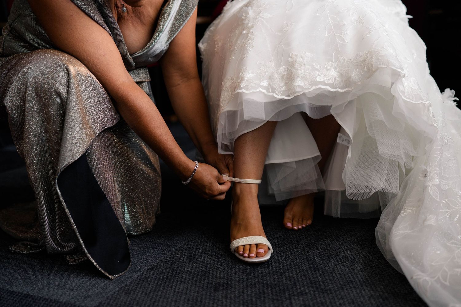 Bridesmaid helping bride fasten white strappy heeled sandal on wedding day preparation.