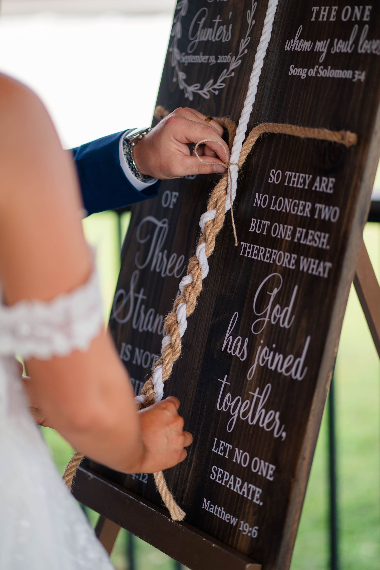 Bride and groom perform cord of three strands unity ceremony at wooden sign display.