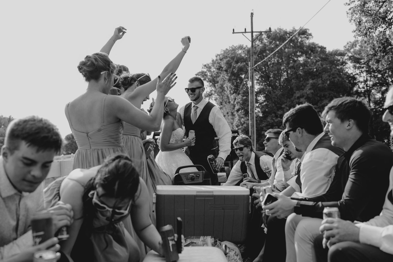 Black and white photo of wedding party celebrating on truck bed with bridesmaids dancing at reception.