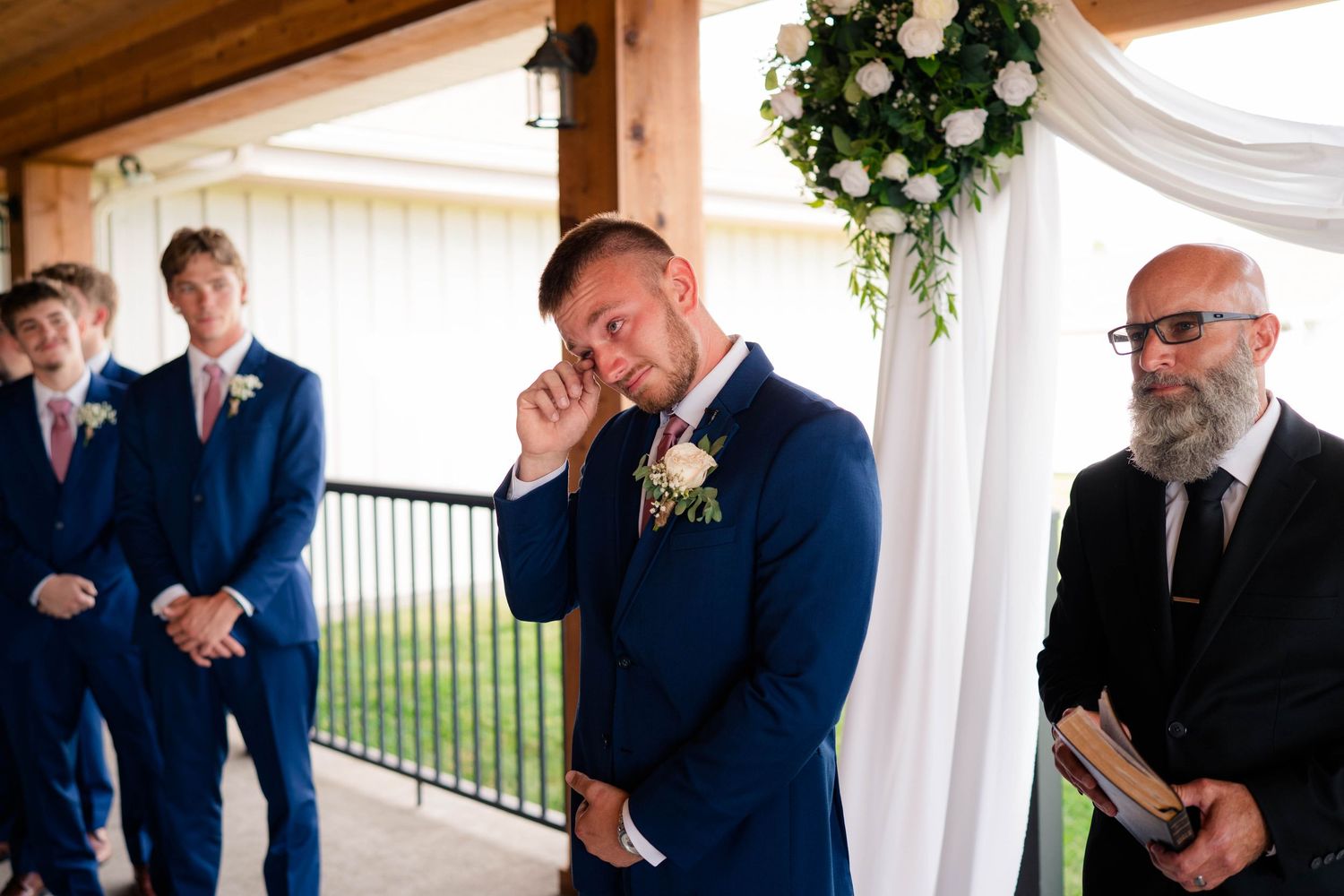 Emotional groom in navy suit wipes tears at the altar as groomsmen and officiant stand nearby during ceremony.