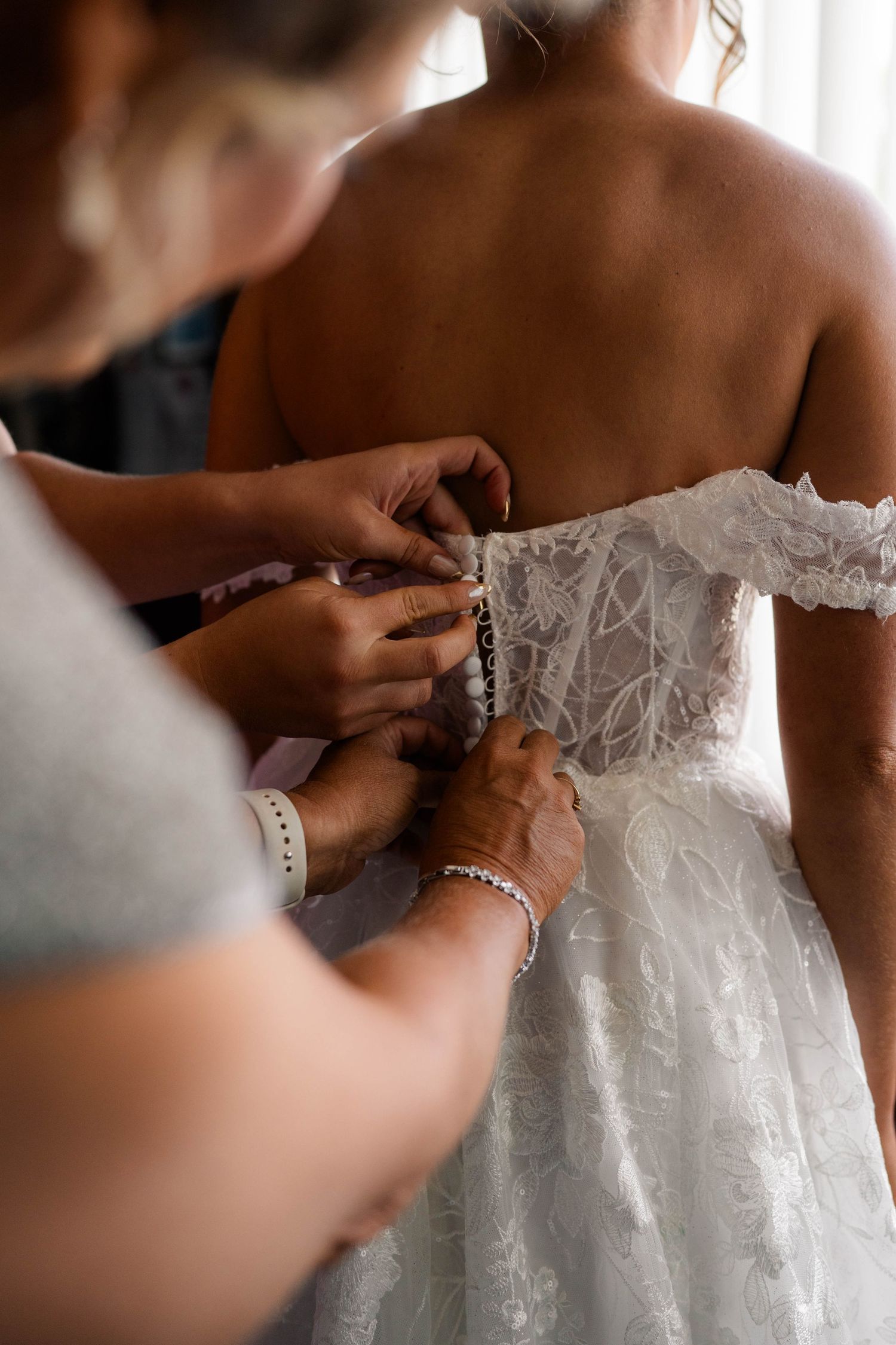 Two women fastening pearl buttons on back of lace bridal gown before wedding ceremony.
