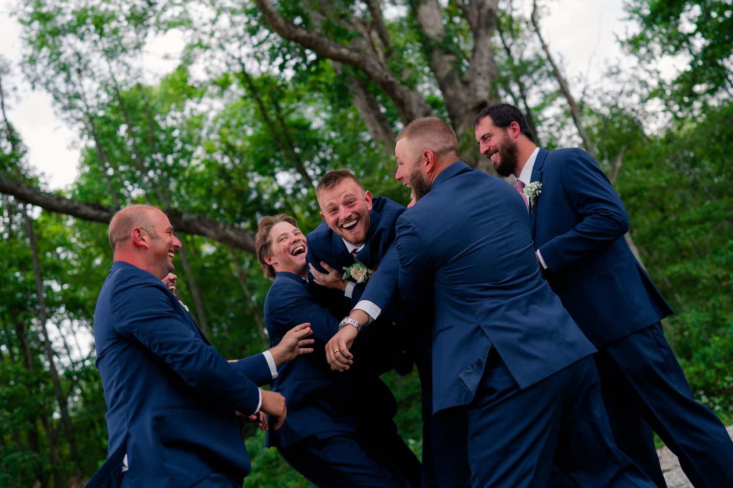 Laughing groomsmen in navy suits playfully lift a fellow groomsman outdoors during a fun wedding party moment.