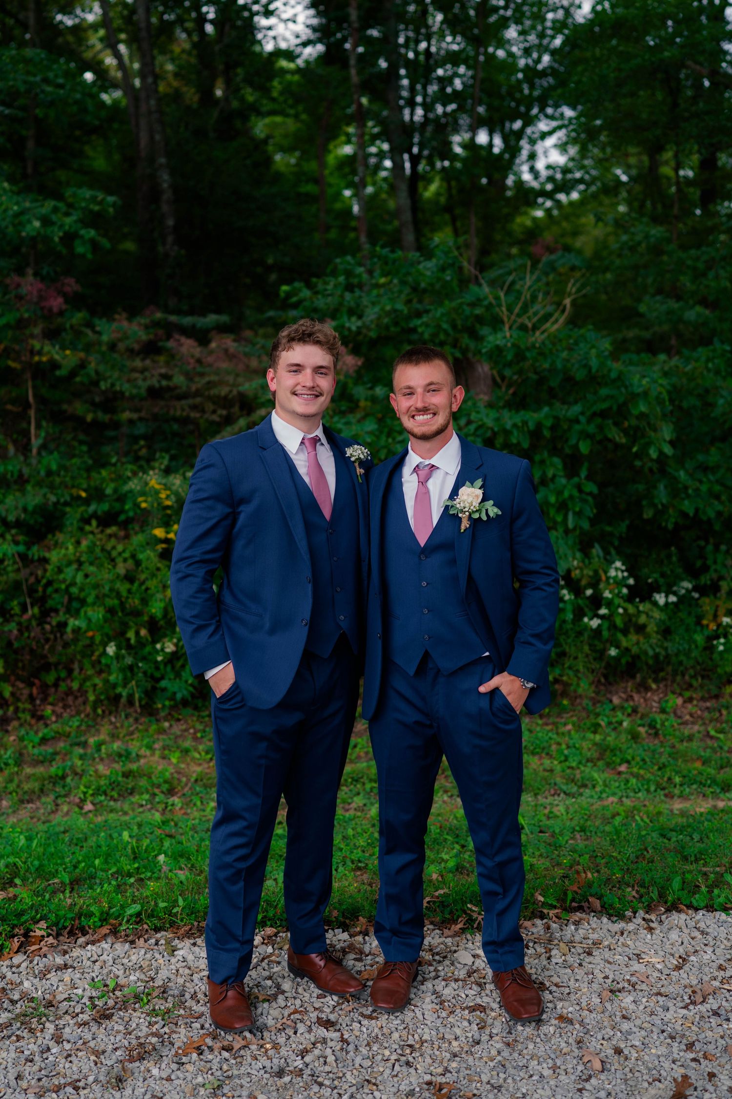 Two groomsmen in navy blue suits with pink ties and boutonnieres posing outdoors at a wedding.