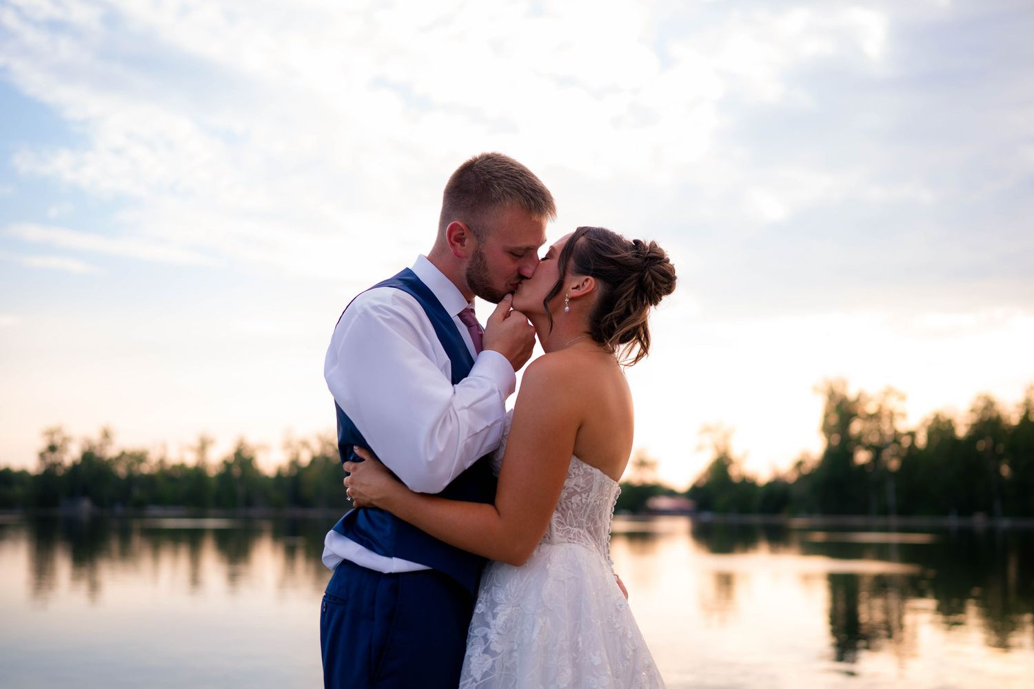 Bride and groom share a romantic moment by a lake at sunset, with the groom in a blue vest and bride in a white gown.
