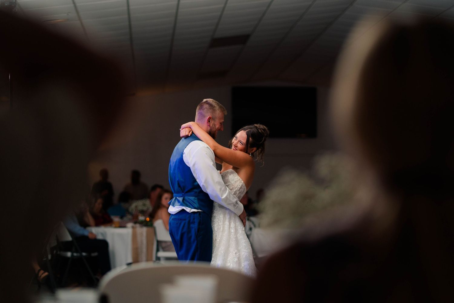 Bride and groom share an intimate first dance in softly lit wedding reception hall with guests watching.