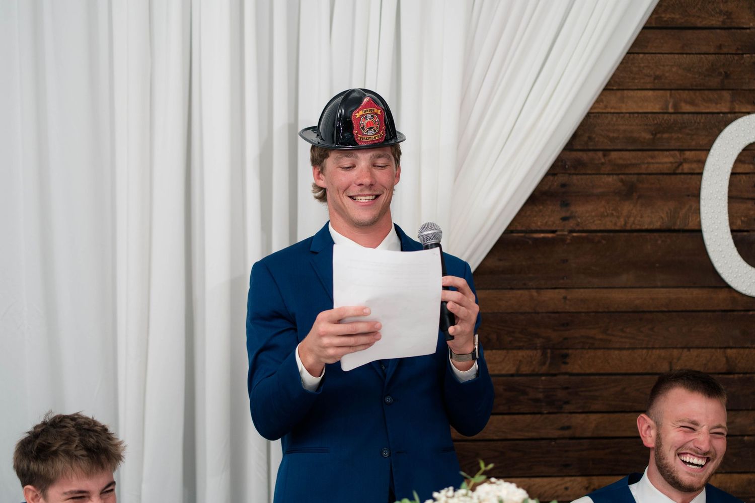 Man in blue suit and firefighter helmet reads speech at wedding reception while guests laugh in background.