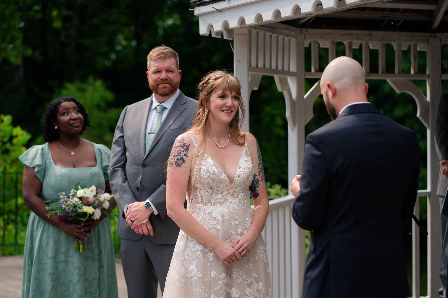 Bride and groom hold hands during outdoor wedding ceremony near white gazebo with bridesmaid holding bouquet.