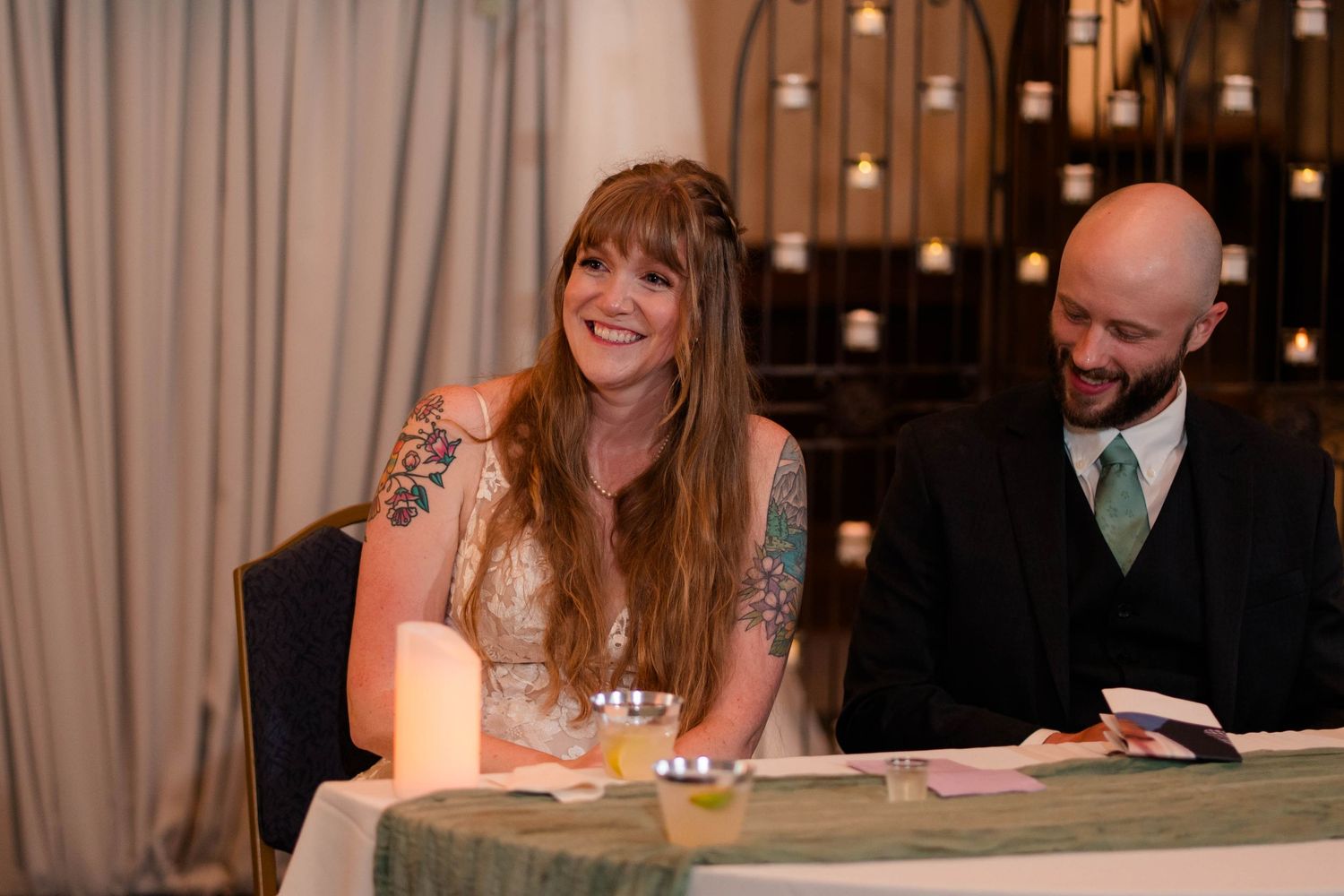 Bride and groom smiling at wedding reception sweetheart table with candles.