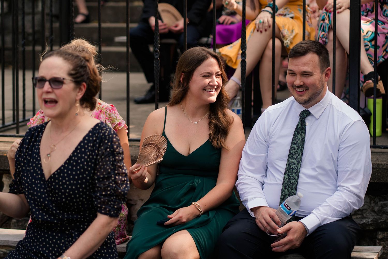 Three wedding guests laughing and smiling while seated outdoors at a summer wedding ceremony.