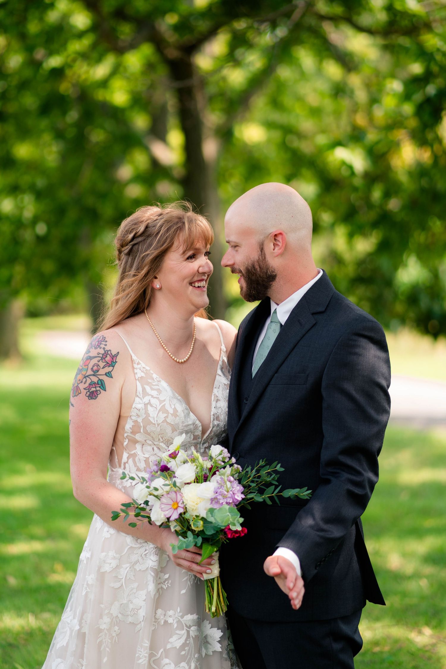 Laughing bride holding a floral bouquet and tattooed arm embraces bald bearded groom in dark suit outdoors.