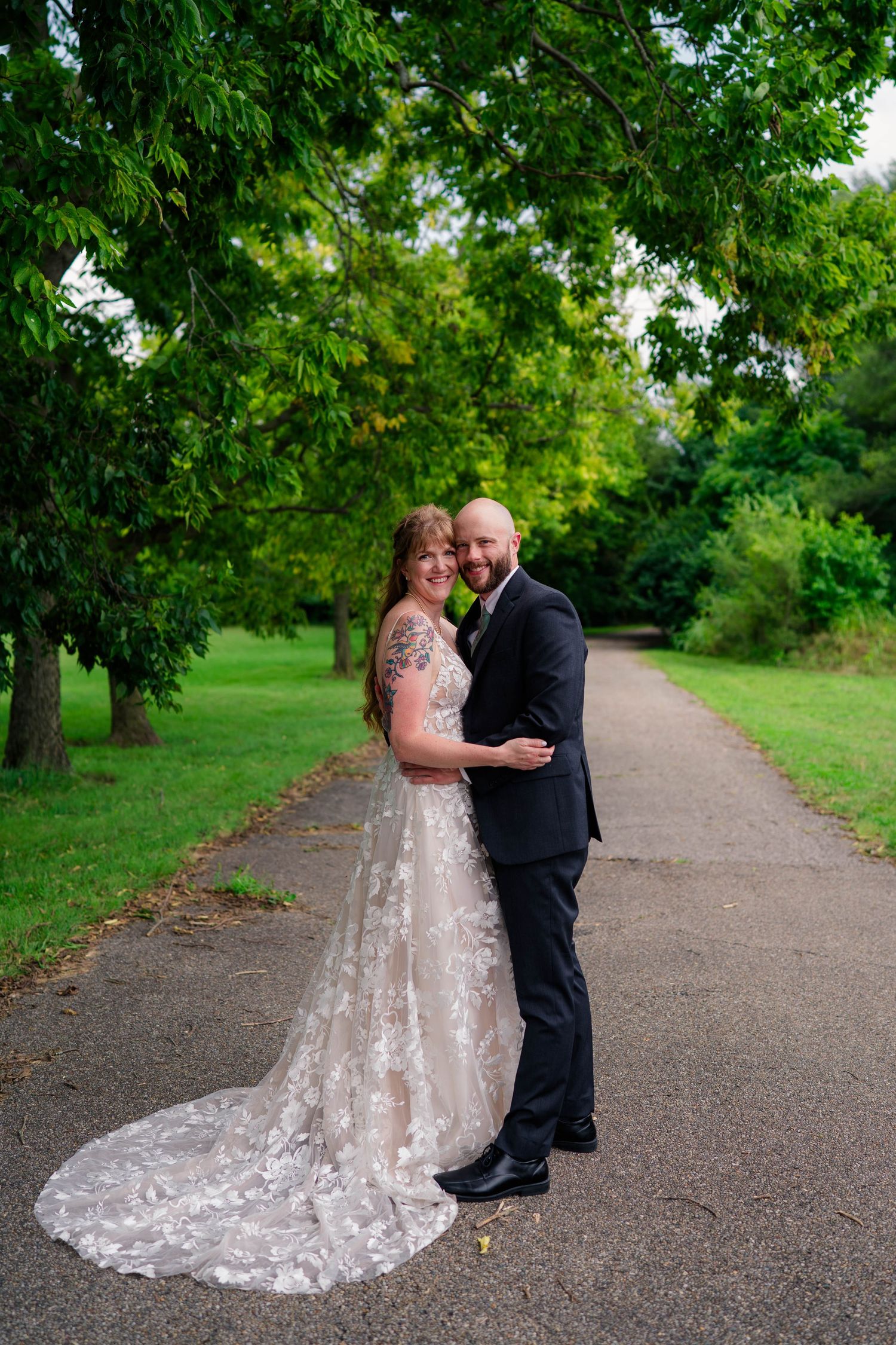 Bride and groom embracing on tree-lined path, she in lace gown with train, he in black suit.