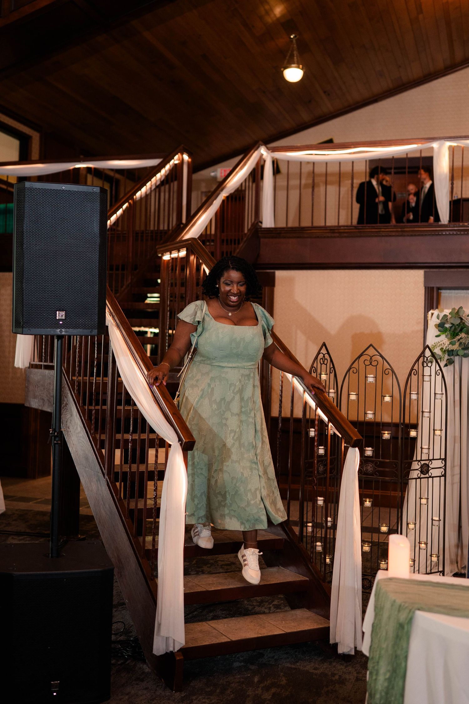 Woman in sage green dress smiles while descending decorated staircase at elegant indoor wedding reception venue.