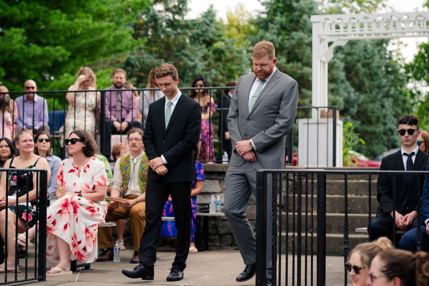 Two groomsmen in dark suits walk toward the ceremony area as wedding guests are seated in the background.
