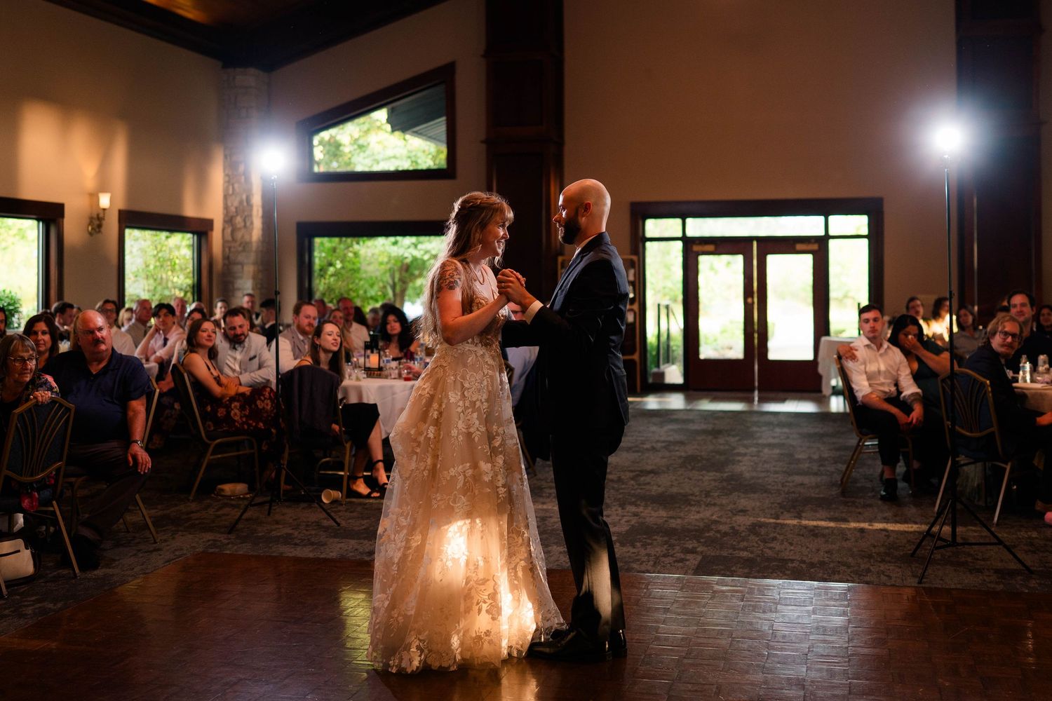 Bride in lace gown and groom share first dance on lit dance floor before seated wedding guests.