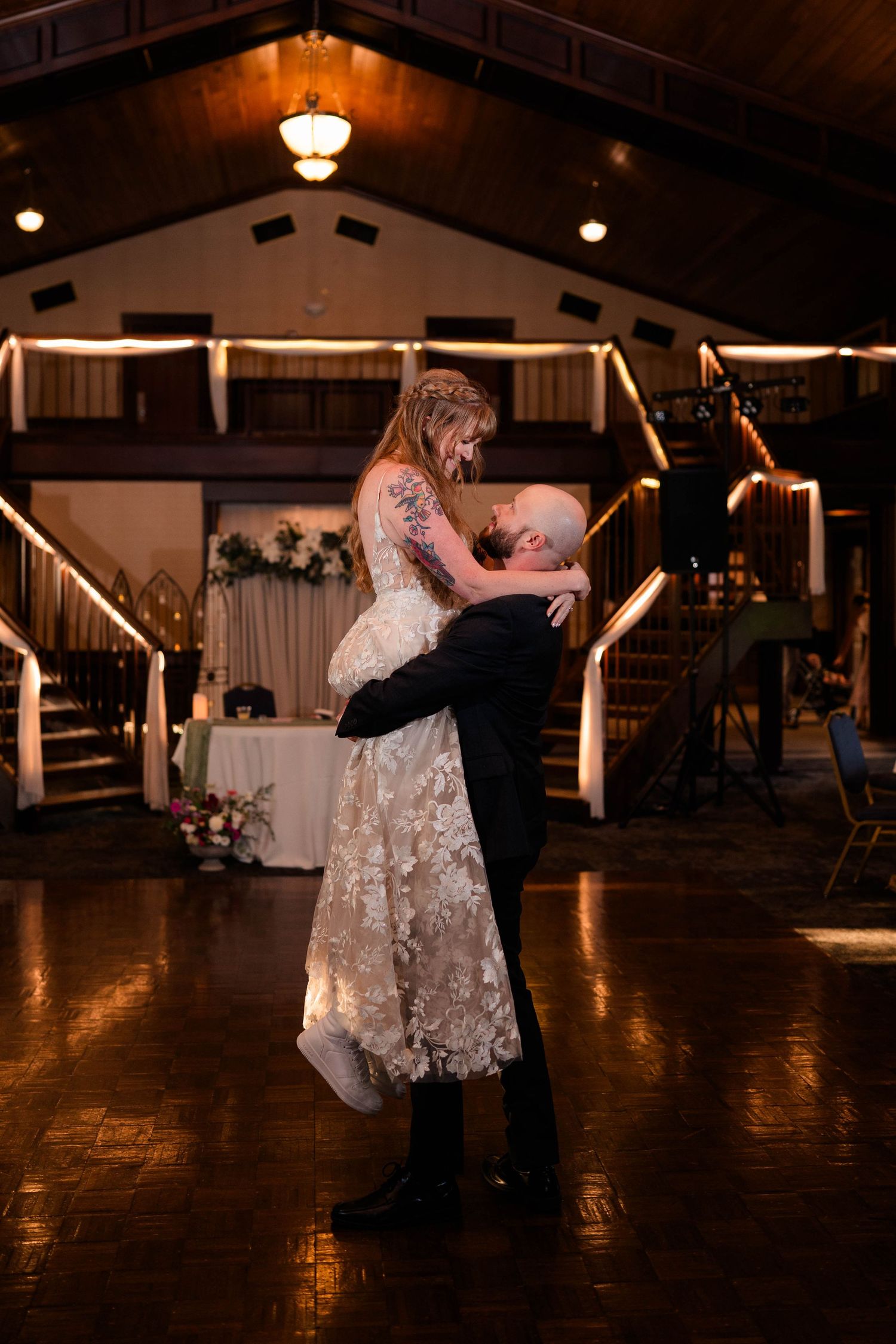 Groom lifting tattooed bride in lace wedding gown during romantic first dance at reception.