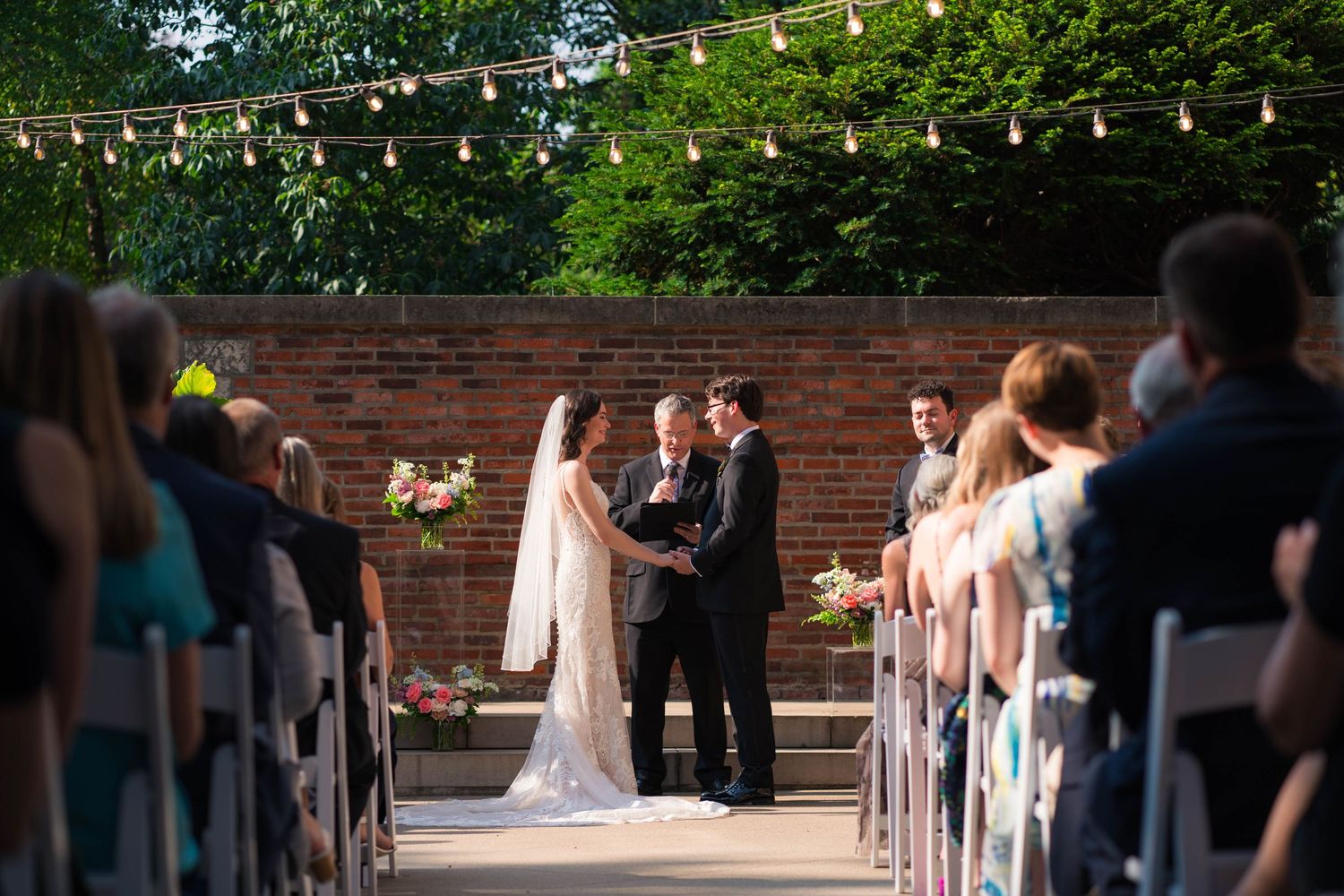 Outdoor wedding ceremony at dusk with string lights, brick wall backdrop, and seated guests in white chairs.